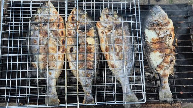 Four whole fish, likely sea bream, sizzling on a charcoal grill inside a wire basket. The skin is golden-brown and charred, with light smoke rising from the hot embers below.