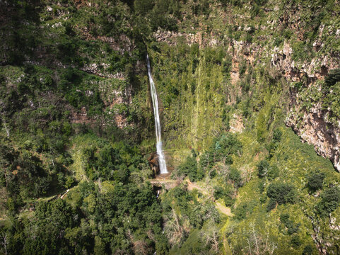 Aerial view of the Cascata do Monte waterfall cascading down a lush green cliffside towards the historic Ponte do Pisao stone bridge in Monte, Madeira, Portugal.