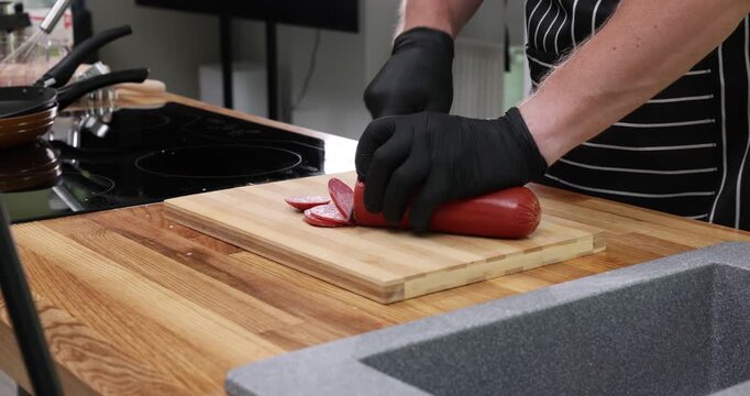 Cook slices sausage on cutting board by sink with stove. Person wearing gloves peels casing trimming meat while setting slices for frying time-lapse
