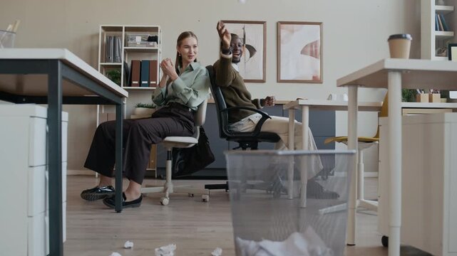 Joyful gen Z Black male and Caucasian female colleagues having fun during break in office, throwing papers into bin from distance