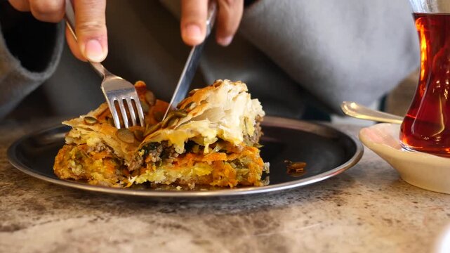 Person cutting savory vegetable pastry with meat and pumpkin seeds on black plate outdoors.