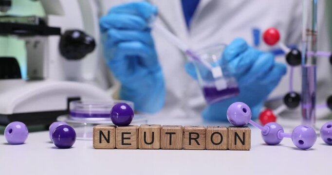 Wooden blocks spell word Neutron near molecule models and lab glassware. Scientist pours solution labelling sample while recording nuclear notes