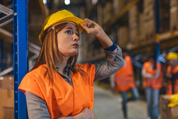 Female Warehouse Worker in Hard Hat and Orange Safety Vest Inspecting Inventory in Industrial...