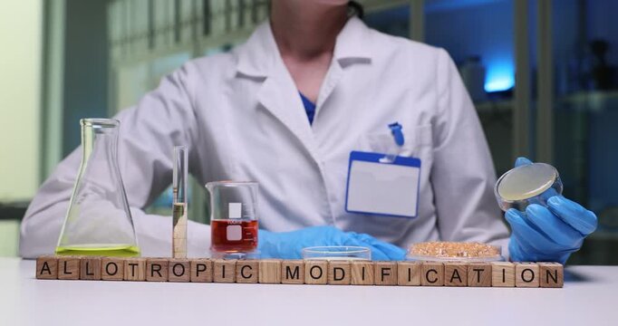 Wooden blocks spell phrase Allotropic Modification near lab flasks and dish. Scientist compares crystal forms recording phase change data carefully
