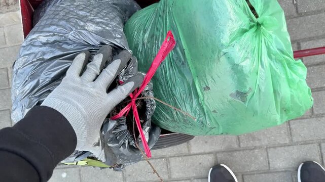A man pushes a red two-wheeled handcart loaded with garbage bags containing bio waste and organic refuse. The scene depicts municipal waste collection and manual transport of household and green waste