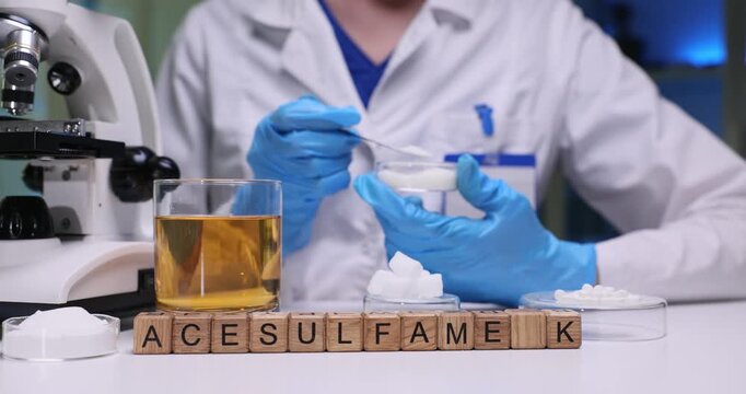 Wooden blocks spell word Acesulfame K near sweetener powder and glassware. Scientist observes additive mixes solution for quality control test