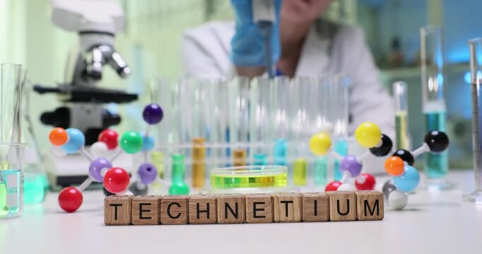 Text Technetium on wooden blocks with molecular structure models on desk. Female chemist adds reagent into test tubes with samples in laboratory