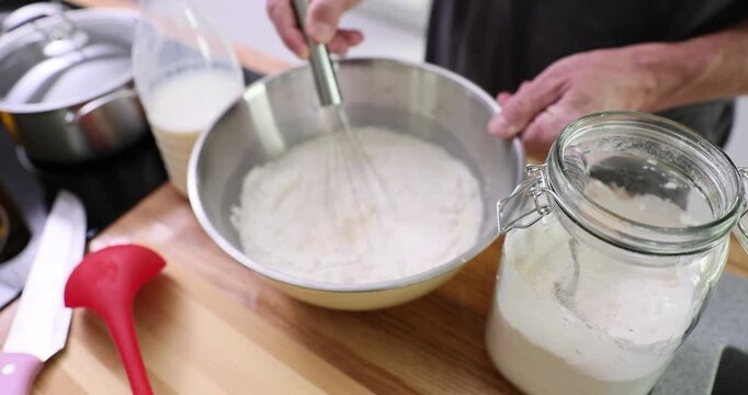 Person whisks milk with flour in steel bowl on wooden counter. Cook smooths liquid quickly checking texture while preparing mixture for pancakes