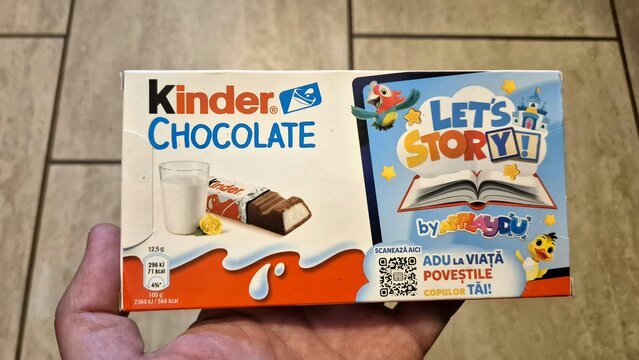A hand holds a 100g box of Kinder Chocolate with a "Let's Story!" promotional design, featuring a glass of milk and a chocolate bar, image taken on April 17, 2026, in Bucharest, Romania.
