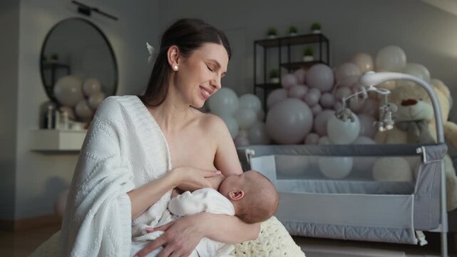 Mother lovingly breastfeeding newborn baby in nursery with balloons and crib. Woman parent child family love parenthood maternity cozy home adorable moment childhood innocence peaceful calm nurturing