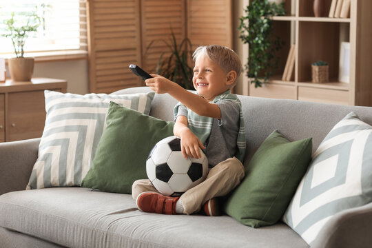 Cute little football player with soccer ball watching TV at home