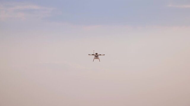 A professional agricultural multi-rotor drone equipped with a camera and antenna slowly hovers and approaches against a slightly cloudy sky, demonstrating modern technology