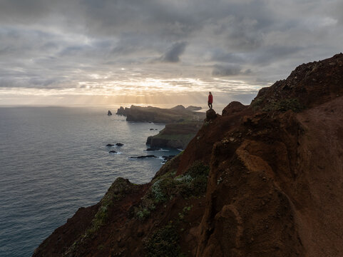 Aerial view of a person in a red jacket standing on the rocky cliffs of Ponta de Sao Lourenco overlooking the Atlantic Ocean under sun rays in Canical, Madeira, Portugal.