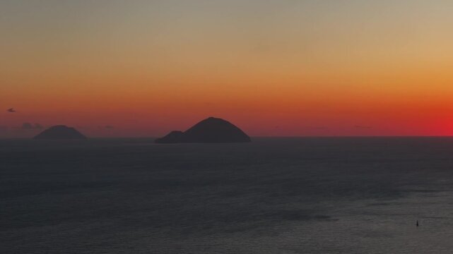 Aerial pan to the right as the sun is halfway below the horizon behind Salina island. Warm glowing sky and volcanic silhouette.