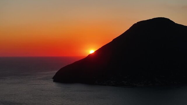Cinematic drone flight moving to the right as the sun sets behind Salina island. Warm orange sky and volcanic silhouette create dramatic Mediterranean sunset atmosphere.