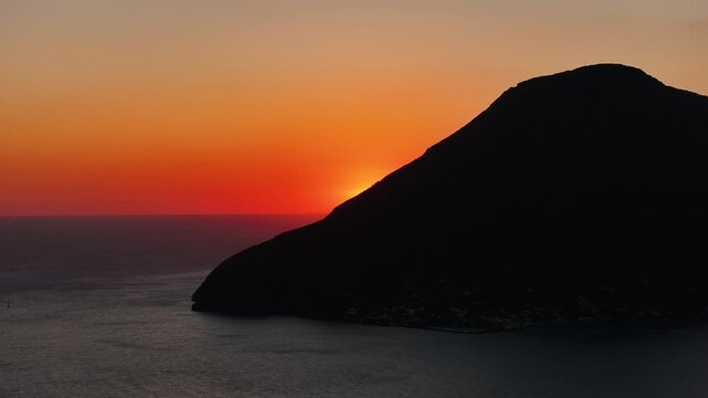 Drone flying left as the sun re-emerges from behind Salina island. Cinematic reverse movement with glowing sunset light over calm sea.