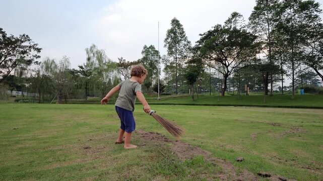 Little boy with short broom playfully sweeps up dry grass clippings in park, enthusiastically imitating adults. Toddler concentrates on his task, but stubborn clumps of grass are rather poorly swept.