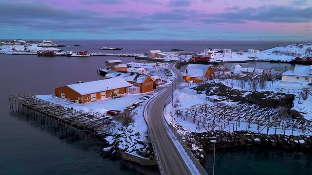 Cinematic drone shot of a snowcovered Norwegian fishing village in Lofoten at sunset. Features traditional rorbu cabins and fish drying racks.