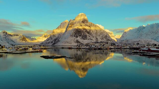 Stunning cinematic drone shot of Reine village with snowcapped mountains reflecting in the calm fjord water during a golden hour sunset in Lofoten, Norway.
