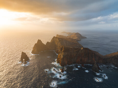 Aerial view of Ponta de Sao Lourenco featuring rugged cliffs, sea stacks, and crashing waves under a golden sunset sky in Canical, Madeira, Portugal.