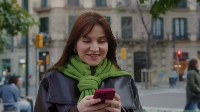 Young smiling woman using smartphone on a modern city street. Social media, messaging, online communication and mobile lifestyle concept. High quality 4k footage