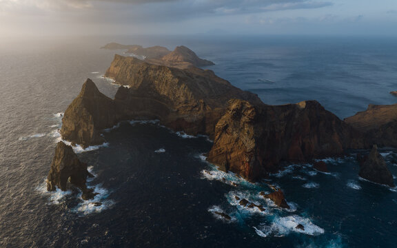 Aerial view of the rugged volcanic cliffs and rocky peninsula of Ponta de Sao Lourenco jutting into the Atlantic Ocean under a dramatic sunset sky in Canical, Madeira, Portugal.