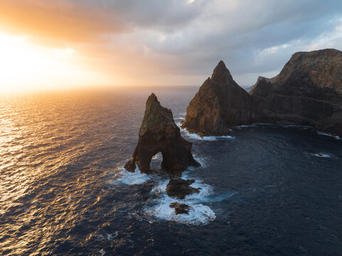 Aerial view of Ponta de Sao Lourenco featuring dramatic sea stacks with a natural stone arch and rugged cliffs under a golden sunset glow in Canical, Madeira, Portugal.
