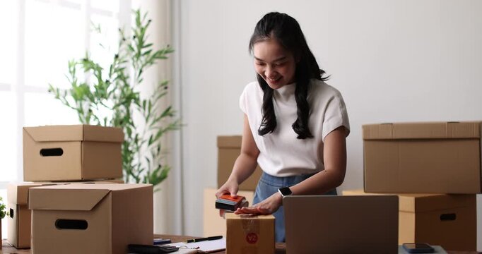 Young Asian woman sitting on floor among shipping boxes, typing on laptop and packing orders while managing her home e-commerce business, focused and efficient in casual apartment setting