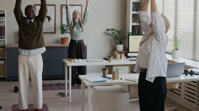 Wide shot of young and senior managers standing on yoga mats in modern office, practicing sun salutation yoga sequence, recharging during workday