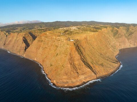 Aerial view of Ponta do Pargo Lighthouse on the edge of steep volcanic cliffs overlooking the Atlantic Ocean with a green plateau and clear blue sky Ponta do Pargo, Madeira, Portugal.