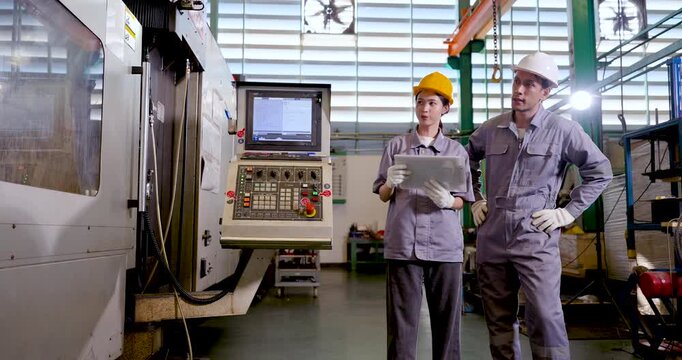 Asian engineer pair stands beside CNC machine with tablet, reviewing production target, machine condition, safety checklist, digital workflow, operational planning in slow motion scene.