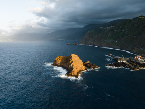 Aerial view of the Ilheu Mole rocky islet and rugged coastline under a dramatic cloudy sky at sunset in Porto Moniz, Madeira, Portugal.