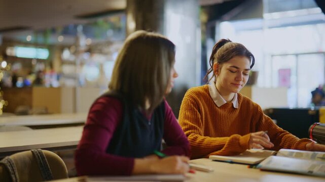 Two young female students are focused on a collaborative study session at a table. They are discussing academic work and writing notes in an educational setting.