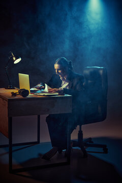 Business woman working late at desk with laptop in dark smoky office lighting. Concept of overtime work, burnout, deadline pressure, productivity stress and corporate lifestyle.
