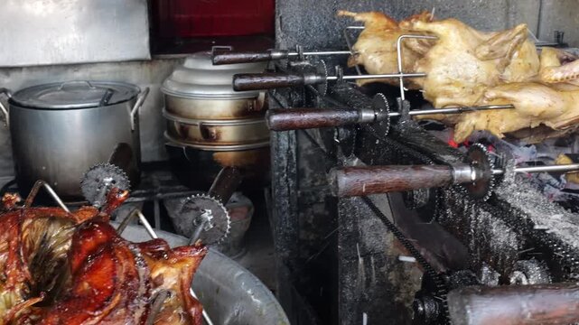 A row of whole poultry carcasses turning on metal rotisserie spits over hot red embers. The skin is golden brown and glistening. The industrial charcoal grill shows visible gears and mechanical parts 