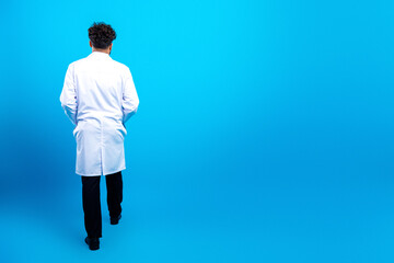 Fototapeta premium Medical professional in a white lab coat walking away on a bright blue studio background, back view of doctor or scientist conveying professionalism and forward movement