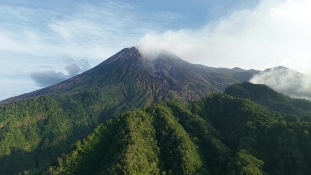 An aerial view of Mount Merapi surrounded by dense forest in Java, Indonesia. A view of volcanic terrain and hills under a clear blue sky.