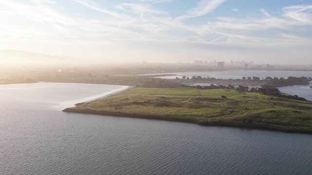 Stunning aerial perspective slowly panning over the lush green landscape of Cesar Chavez Park and Albany Beach in Berkeley, California, with the hazy city skyline visible in the distance at sunrise