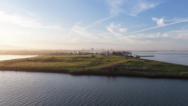 Morning sunlight illuminating the grassy peninsula of Cesar Chavez Park and Albany Beach, with the city of Berkeley visible in the hazy distance across the calm waters of the bay
