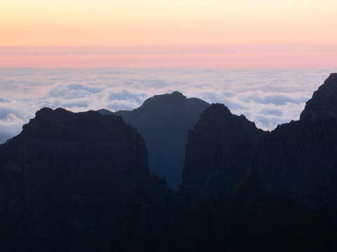 Aerial view of Pico do Arieiro mountain peaks rising above a thick sea of clouds during a colorful sunset in Curral das Freiras, Madeira, Portugal.