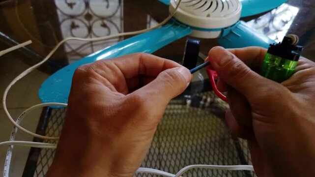 Close up shot of Hands holding a spliced wire wrapped with smooth black electrical tape. Red scissors and blue fan parts on a glass table. DIY home appliance repair.