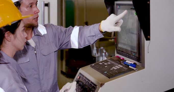 Caucasian man engineer points at CNC display while Asian woman technician checks screen content, supporting machine setting review and production control in workshop, slow motion