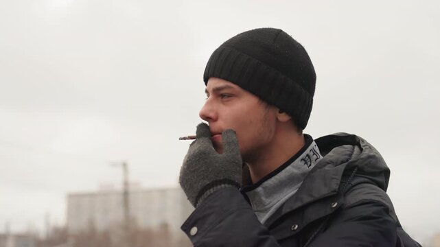 Young white man smoking cigarette outdoors, bundled beanie winter coat gloves, overcast city skyline blurred in background, breath visible in cold air, contemplative profile shifting to direct gaze,