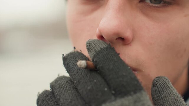 Contemplative youth enjoying cigarette. Thoughtful young person inhales cigarette smoke peacefully. Youthful individual with gloves and beard calmly exhaling smoke in urban setting
