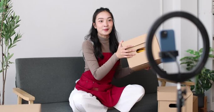 Asian woman livestreaming fashion from a cozy home studio, promoting her small ecommerce boutique on social media using smartphone and ring light to showcase clothes and connect with customers
