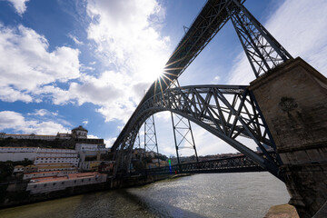 Fototapeta premium Steel arch bridge over a river with city buildings and sun in a blue sky.