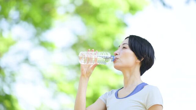 新緑の屋外で水分補給をする女性の横顔ポートレート　ウォーキングやランニングの運動やスポーツの間にペットボトルの水を飲むクローズアップ　春夏の半袖の動画