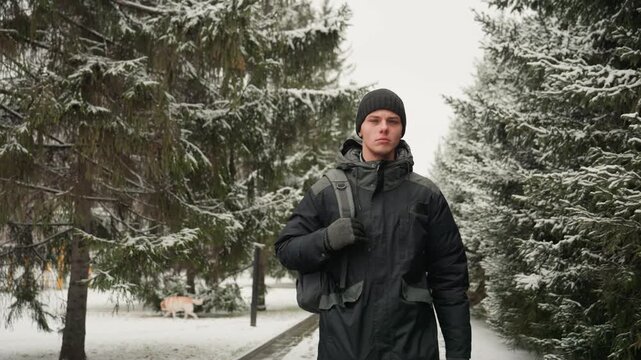 Winter park walkway closeup of pedestrian, young man in black jacket and beanie carrying backpack, steady stride toward camera, snow on evergreen branches, brisk commute mood, crisp cold light