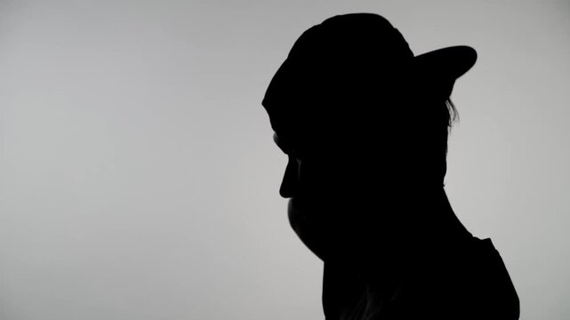 An anonymous silhouette of a man against a white wall puts his cap on backwards, shakes his head and takes it off