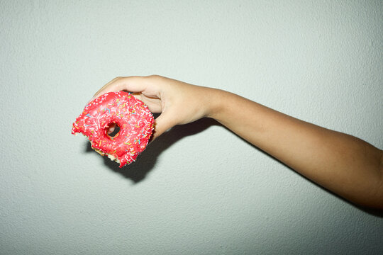 Close up of adult hand holding bitten pink frosted donut with sprinkles against plain wall, suggesting indulgence and craving. Useful for junk food, sugar consumption, diet messaging